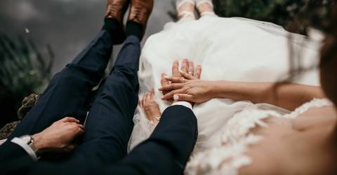 Brui met bruidegom zitten langs een waterkant met bungelende benen boven het water met de handen op elkaar waar een trouwring te zien is